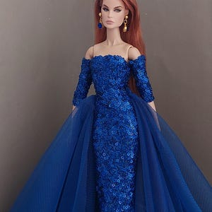 May include: A fashion doll with long auburn hair wearing an off-the-shoulder, royal blue sequined gown with three-quarter length sleeves and a voluminous tulle overskirt. The doll is accessorized with gold-toned drop earrings featuring blue stones.