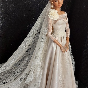 Könnte beinhalten: Eine Porzellanpuppe in einem weißen Spitzen-Hochzeitskleid mit langem Schleier. Die Puppe hat blonde Haare und trägt Perlenohrringe.