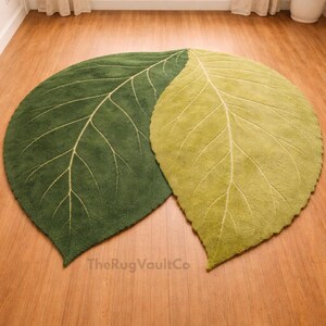 May include: Two leaf-shaped rugs in shades of green and yellow-green rest on a wooden floor. The rugs have a textured surface with cream-colored vein detailing. The Rug Vault Co. is printed on the bottom.