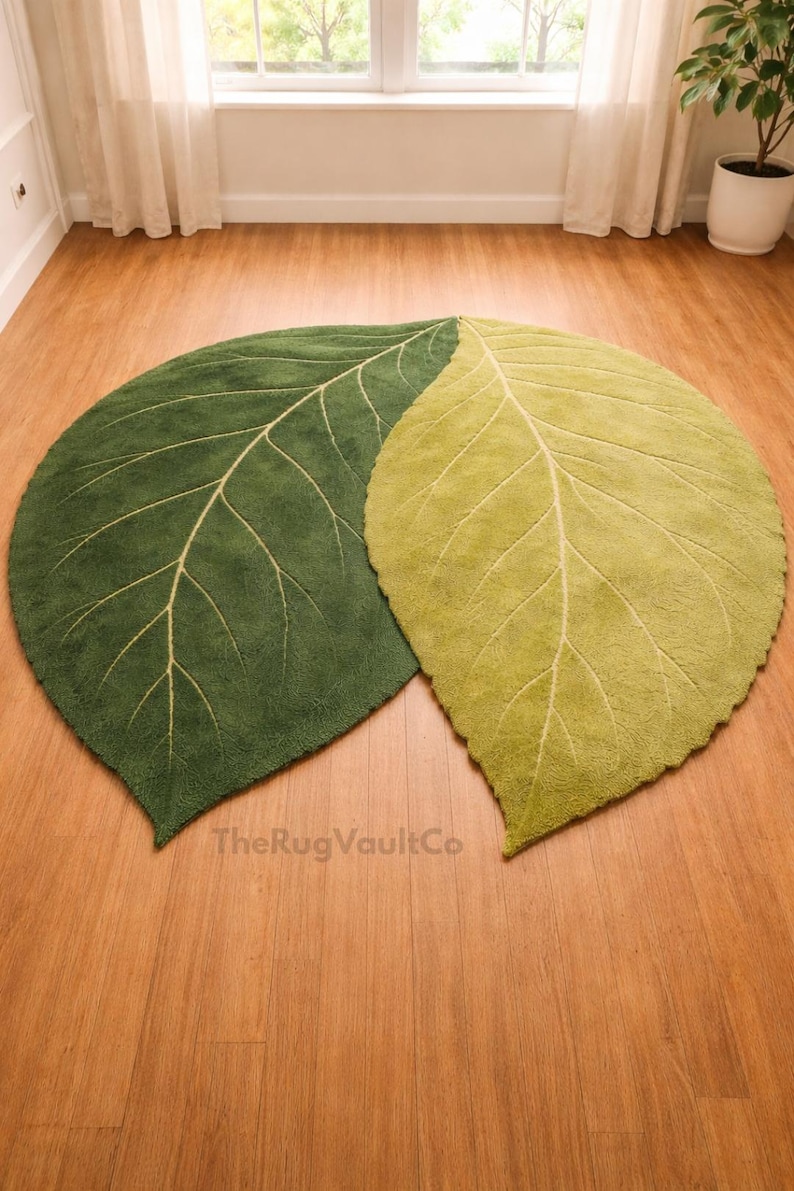 May include: Two leaf-shaped rugs in shades of green and yellow-green rest on a wooden floor. The rugs have a textured surface with cream-colored vein detailing. The Rug Vault Co. is printed on the bottom.