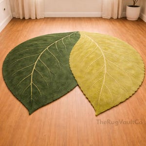 May include: Two leaf-shaped rugs in shades of green and yellow-green rest on a wooden floor. The rugs have detailed vein patterns in a lighter color, mimicking real leaves. The rugs are placed in front of a window with sheer curtains.