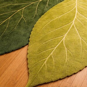 May include: Two leaf-shaped rugs in shades of green. The rugs have a textured surface and detailed vein patterns in a lighter green color. The rugs are placed on a wooden floor.