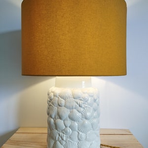 Puede incluir: Una lámpara de mesa de cerámica blanca con una base con forma de conchas. La lámpara tiene una pantalla de tela de color amarillo mostaza.