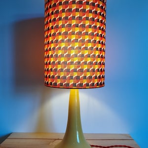Puede incluir: Una lámpara de mesa vintage con una base de cerámica amarilla y una pantalla cilíndrica con un patrón geométrico en tonos naranja, marrón y negro.