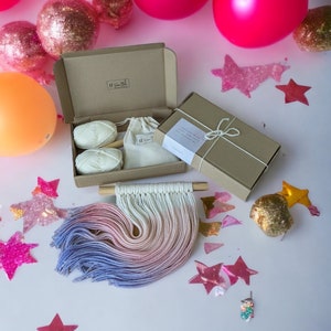May include: A DIY macrame wall hanging kit with two balls of yarn, a wooden dowel, and a small bag of instructions. The kit is packaged in a brown cardboard box with a white label that says "Created with love for your home." The kit is surrounded by pink and gold star-shaped confetti and pink balloons.
