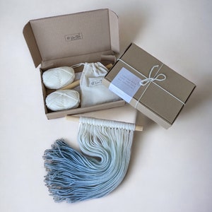Puede incluir: Un kit de manualidades con dos ovillos de hilo blanco, una varilla de madera y una bolsa pequeña. El kit está empaquetado en una caja de cartón marrón con el texto "Crafted with love for your DIY adventure!" en una etiqueta blanca.
