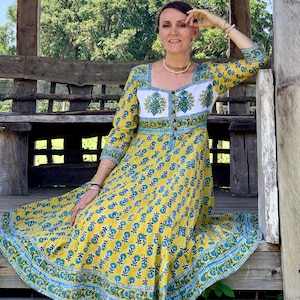 May include: A long, yellow dress with a blue floral pattern and white bodice. The dress has long sleeves and a square neckline. The woman is sitting on a wooden bench in an outdoor setting. The dress is a bohemian style.