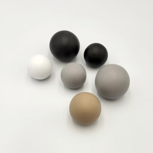 Puede incluir: Un conjunto de seis esferas de madera en varios colores, incluyendo negro, blanco, gris y marrón. Las esferas están dispuestas en un semicírculo sobre un fondo blanco.