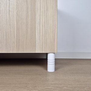 Könnte beinhalten: Ausschnitt eines hellen Holzschranks mit einem weißen zylindrischen Bein. Das Bein hat drei horizontale Rillen. Der Schrank steht auf einem hellbraunen Holzboden, mit einer weißen Wand im Hintergrund.