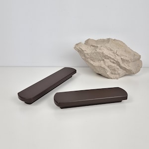Op de afbeelding: Twee donkerbruine kastgrepen met afgeronde uiteinden worden getoond op een wit oppervlak. Een getextureerde, lichtgrijze rots bevindt zich op de achtergrond. De grepen zijn rechthoekig en lijken van hout of een vergelijkbaar materiaal te zijn.