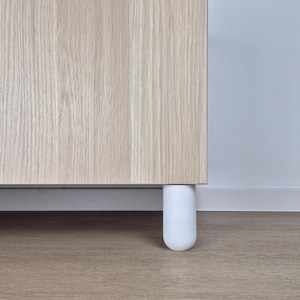 Könnte beinhalten: Nahaufnahme eines hellen Holzschranks mit einem weißen, abgerundeten Bein. Der Schrank hat eine natürliche Holzmaserung. Das Bein ist zylindrisch und scheint aus einem festen Material zu bestehen.