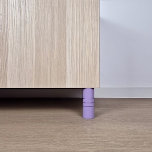 Op de afbeelding: Een licht houten kast met zichtbare houtnerf. De kast staat op korte, cilindrische lavendelkleurige poten, elk met drie horizontale groeven. De kast staat tegen een witte muur en een lichtbruine vloer.