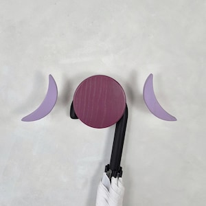 May include: A wall-mounted umbrella holder with a circular, dark purple wooden base and two crescent-shaped, light purple hooks. The umbrella handle is black, and the umbrella itself is white. The design is minimalist and modern.