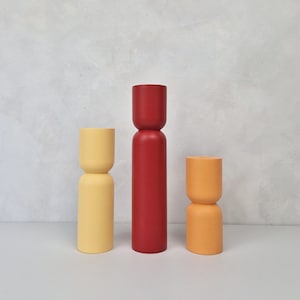 May include: Three wooden decorative objects in yellow, red, and orange. The objects have a cylindrical shape with a rounded top and bottom. The background is a light grey wall.