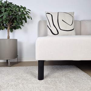 Pode incluir: Um sofá creme com pernas pretas e uma almofada com um design abstrato preto sobre um fundo creme. À esquerda, há uma planta num vaso cinza e, em frente, um tapete branco. O sofá tem um design moderno.
