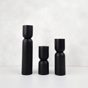 May include: Three black wooden candle holders of varying heights. The candle holders have a unique hourglass shape with a wider middle section.