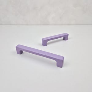 May include: Two lavender cabinet pulls, one longer than the other, with a rectangular shape and rounded edges.