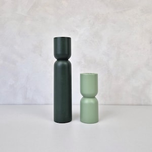 Könnte beinhalten: Zwei Kerzenhalter aus Holz im minimalistischen Stil. Einer ist hoch und dunkelgrün, der andere ist kürzer und hellgrün. Die Kerzenhalter haben eine zylindrische Form mit abgerundeten Ober- und Unterseiten. Sie werden auf einer weißen Oberfläche vor einem neutralen Hintergrund präsentiert.