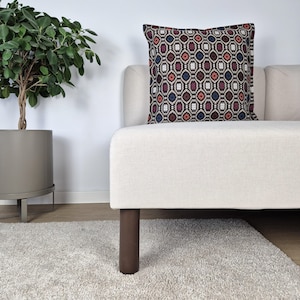 May include: A patterned cushion in shades of red, blue, and beige sits on a cream-coloured sofa. The cushion features a repeating geometric design. The sofa has dark brown wooden legs. The sofa is placed on a light-coloured rug.