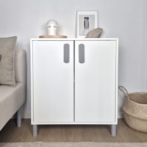 May include: A white cabinet with two doors and gray handles. The cabinet sits on short gray legs. A small white lamp, a framed picture, and a decorative stone are on top of the cabinet. A woven basket is to the right.