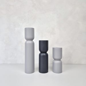 May include: Three modern decorative wooden objects in varying sizes and shades of gray and black. The objects have a unique hourglass or stacked cylinder design. The tallest is a light gray, the middle is dark gray, and the smallest is a lighter gray.