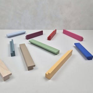 Puede incluir: Una colección de tiradores de armario de madera de colores en varios tonos, incluyendo azul, verde, rosa y amarillo. Los tiradores son rectangulares con un pequeño tirador saliente. El fondo es una superficie lisa y clara.