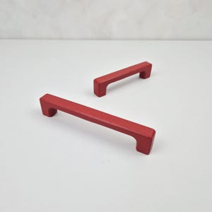 May include: Two red cabinet door pulls with a rectangular shape and a slight curve in the middle.