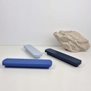 May include: Three rectangular cabinet pulls in shades of blue and gray are arranged on a white surface. The pulls have rounded edges and a matte finish. A textured, light-colored rock is in the background.