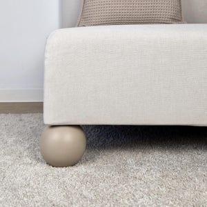 May include: Close-up of a beige upholstered bench with a rounded, light brown leg. The bench sits on a light gray carpet. A beige waffle-weave pillow is visible in the background.