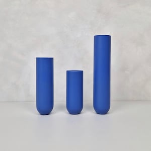 Può includere: Tre vasi cilindrici blu cobalto di altezze variabili. I vasi hanno una finitura liscia e opaca e fondi arrotondati. Sono disposti in fila su uno sfondo grigio neutro, creando un'estetica minimalista. Il vaso più alto è alto circa 30 cm.