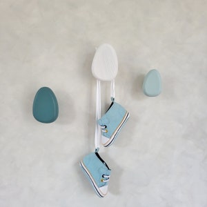 Può includere: Tre ganci in ceramica blu chiaro a forma di ciottoli, un gancio in ceramica bianca a forma di ciottolo e due scarpe da ginnastica per bambini di colore blu chiaro appese al gancio bianco.