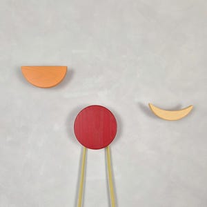 Puede incluir: Tres perchas de pared de madera con un diseño minimalista. Las perchas tienen forma de semicírculo, círculo y media luna. Las perchas están pintadas en naranja, rojo y amarillo, con cuerdas doradas.