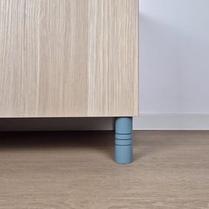 May include: A section of a light wood cabinet with a pale blue cylindrical leg. The leg has three horizontal grooves. The cabinet is set on a light brown wood floor.