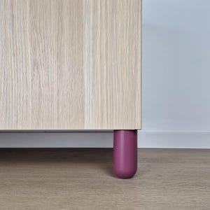 May include: A light wood cabinet section with a single, rounded, dark purple leg. The cabinet has a natural wood grain pattern. The cylindrical leg provides a contrast against the neutral tones of the cabinet.