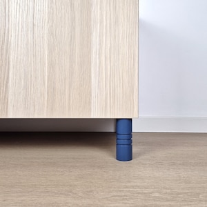 Könnte beinhalten: Ein hellfarbener Holzschrank mit einem blauen zylindrischen Bein. Der Schrank hat eine natürliche Holzmaserung. Das blaue Bein ist einfarbig und hat drei horizontale Rillen. Der Schrank steht auf einem hellfarbenen Holzboden.