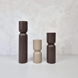 Puede incluir: Tres objetos decorativos de madera de diferentes alturas y colores. El más alto es marrón oscuro, el del medio es beige y el más corto es marrón oscuro. Los objetos tienen forma cilíndrica con la parte superior redondeada.