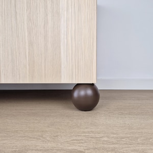 Könnte beinhalten: Ein Teil eines hellfarbenen Holzschranks mit einem dunkelbraunen, kugelförmigen Fuß. Der Schrank steht vor einer neutralen Wand und ruht auf einem hellbraunen Boden. Der Fuß bietet Halt und eine moderne Ästhetik.
