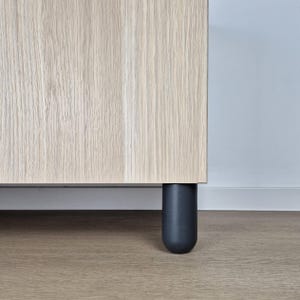 Op de afbeelding: Close-up van een lichtgekleurde houten kast met een donkergrijze, afgeronde poot. De kast heeft een verticaal houtnerfpatroon. De poot is cilindrisch met een afgeronde basis.