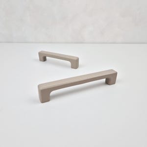May include: Two beige cabinet pulls with a rectangular shape. The pulls are made of a smooth, matte material.