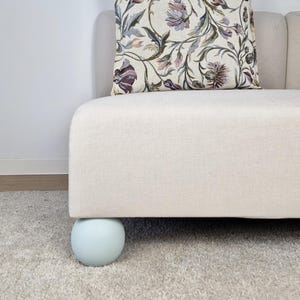 May include: A light-colored sofa with a floral patterned pillow. The sofa has a rounded, light blue leg. The pillow features a design with purple, green, and beige tones. The sofa is placed on a light gray carpet.