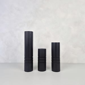 May include: Three black cylindrical decorative objects of varying heights. Each has horizontal grooves near the base. The objects are arranged on a white surface against a light grey textured background. The tallest object is approximately 25 cm tall.