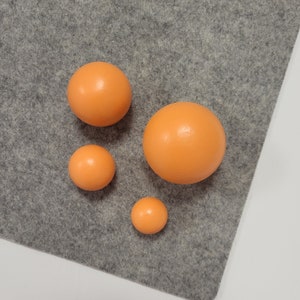 Puede incluir: Cuatro esferas naranjas de diferentes tamaños dispuestas sobre una superficie de fieltro gris. Las esferas tienen un acabado liso y mate. El fondo es de color gris sólido.