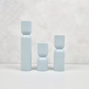 May include: Three light blue ceramic candle holders of varying heights. The candle holders are cylindrical with a rounded top and a slightly wider base.