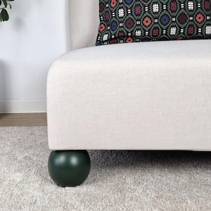 Può includere: Una sezione di divano color crema con una gamba sferica verde scuro. Un cuscino fantasia con un design geometrico nei toni del rosso, blu e verde poggia sul divano. Il divano è su un tappeto chiaro.