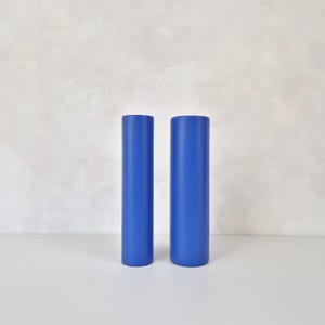 Pode incluir: Dois objetos cilíndricos de uma cor azul vibrante. Cada peça apresenta um acabamento liso e sólido com um topo plano e uma base arredondada. Os dois itens são idênticos em forma e tamanho, posicionados lado a lado como um par.