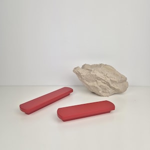 May include: Two red rectangular containers with rounded edges are displayed on a white surface. A textured, light-colored rock sits in the background. The containers appear to be made of a solid material.