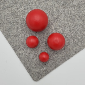 May include: Four red wooden spheres of different sizes are displayed on a grey felt surface. The spheres vary in size, creating a visual gradient. The red colour is uniform across all spheres.