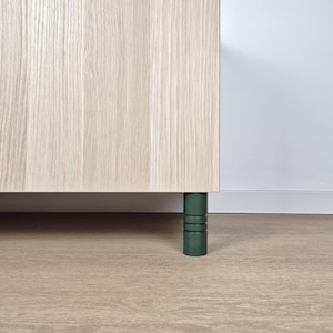 Op de afbeelding: Een gedeelte van een lichtgekleurde houten kast met een groene, cilindrische poot. De poot heeft drie horizontale groeven. De kast staat op een lichtbruine houten vloer, met een witte muur op de achtergrond.
