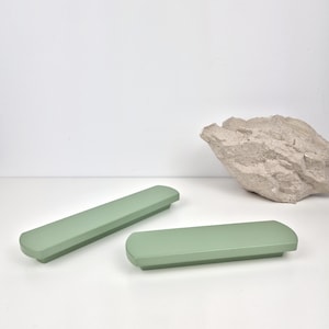Può includere: Due maniglie per armadietti verde salvia con bordi arrotondati sono esposte su una superficie bianca. Sullo sfondo c'è una grande roccia ruvida di colore chiaro. Le maniglie sono rettangolari e sembrano essere in legno o materiale simile.