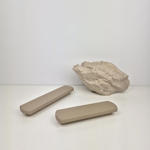 May include: Two taupe-colored, rectangular cabinet pulls with rounded ends are displayed. A light-colored, textured rock sits in the background. The cabinet pulls are made of a solid material and are designed for furniture.